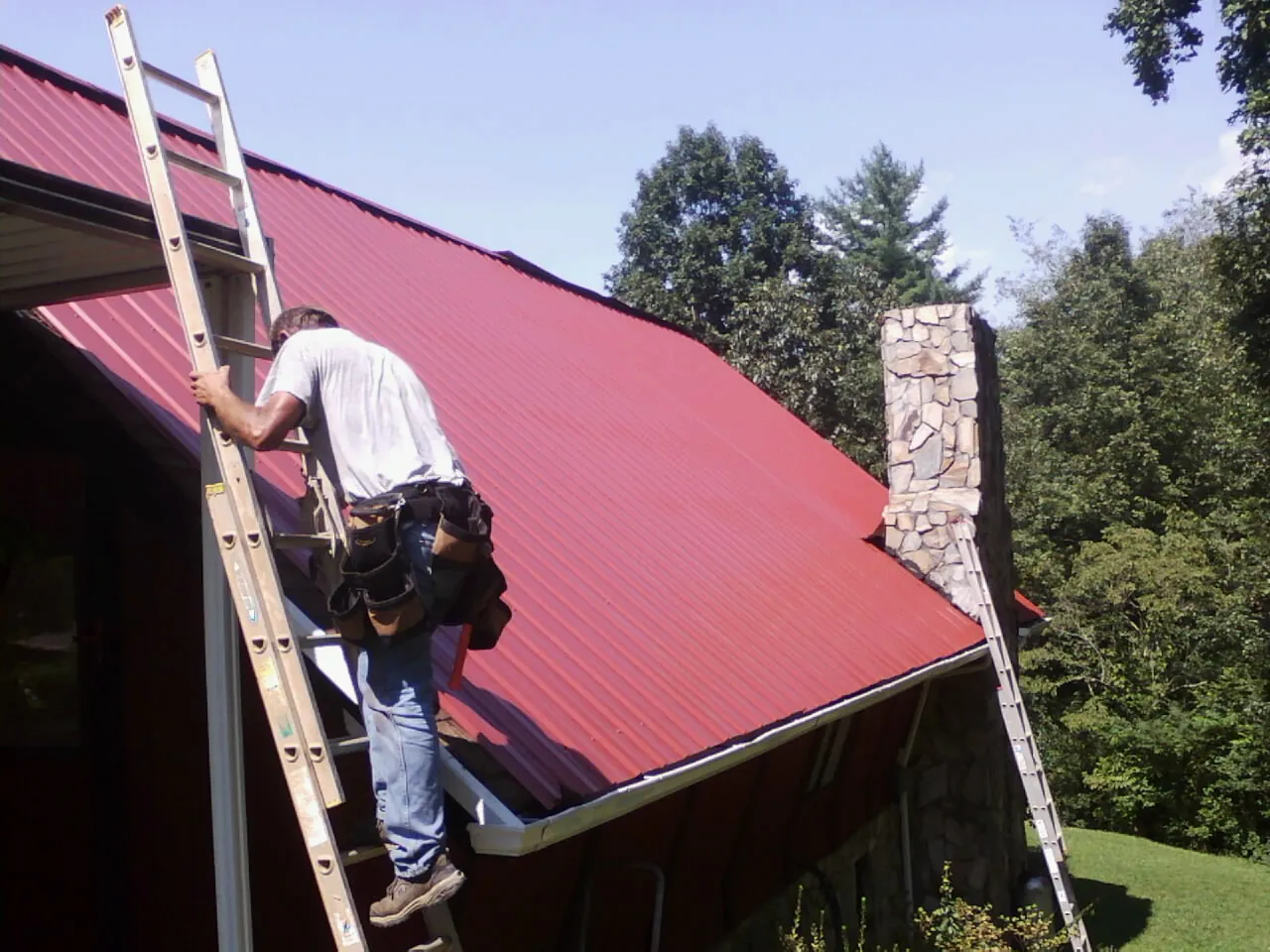 Roofer on a ladder beside a red metal roof and stone chimney — Soffit & Fascia Repair by Roofing Brothers in Inman